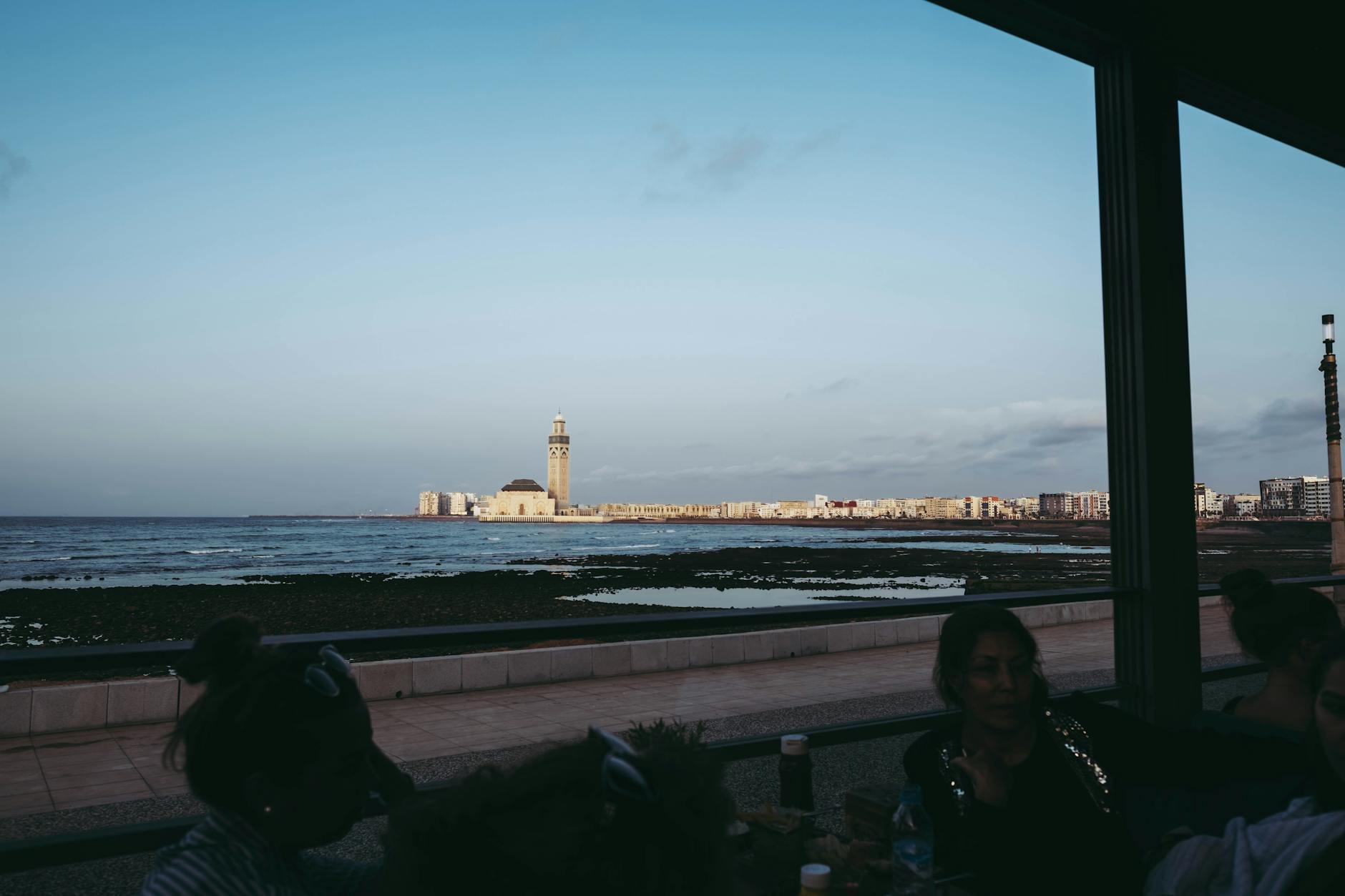 Scenic view of Hassan II Mosque from the Casablanca coastline during daytime with people in a café foreground.