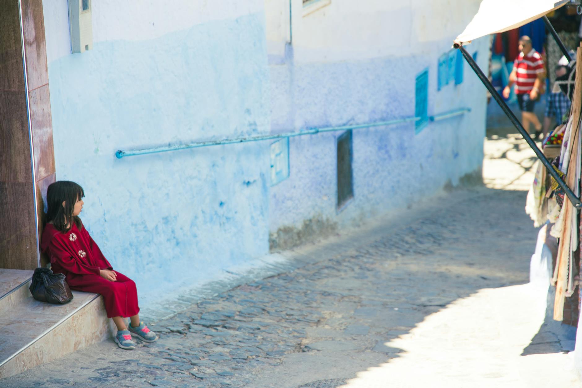A child in red attire sits by a blue wall in a bustling Moroccan street.